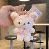 A pink cherry blossom plush bear keychain is held by a hand against a blurred indoor background.