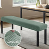 A green stretch jacquard bench cover placed on a wooden table with a glass vase in a bright room.