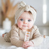 A baby wearing a beige knitted sweater is holding a wooden rain rattle sensory toy.