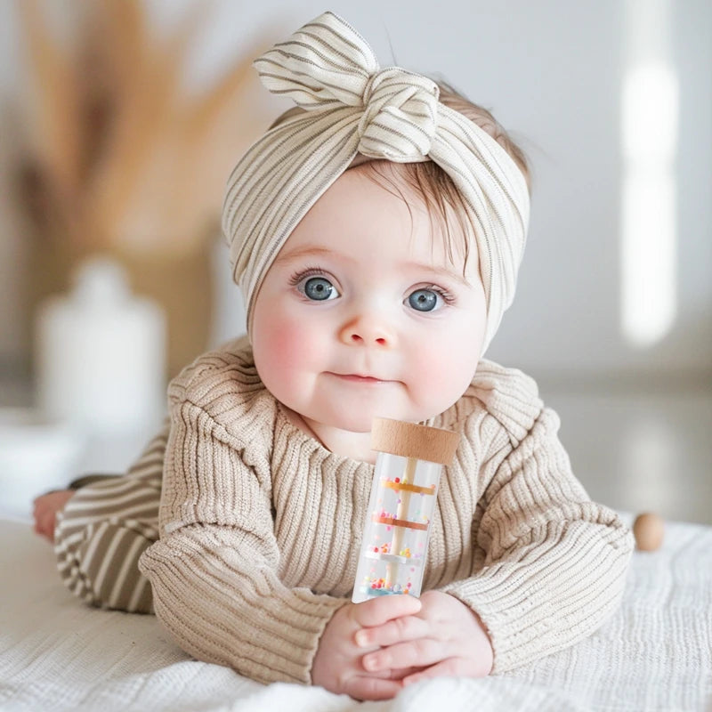 A baby wearing a beige knitted sweater is holding a wooden rain rattle sensory toy.