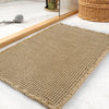 A beige waffle texture bath mat is displayed on a white marble surface next to wooden slats and a wicker basket.