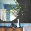 A decorative vase with green branches sits on a dark surface in front of a textured gray wall and a blue painted wainscoting.