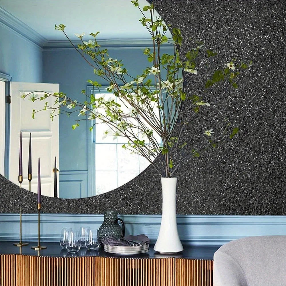 A decorative vase with green branches sits on a dark surface in front of a textured gray wall and a blue painted wainscoting.