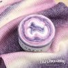 A roll of colorful gradient cake yarn is displayed on a purple fabric background.