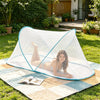 A woman relaxing on a light blue pop-up mosquito net spread on an outdoor mat in a grassy yard.