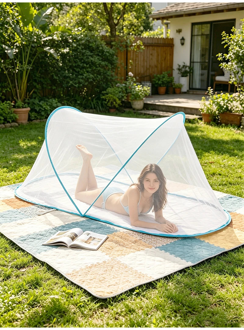 A woman relaxing on a light blue pop-up mosquito net spread on an outdoor mat in a grassy yard.