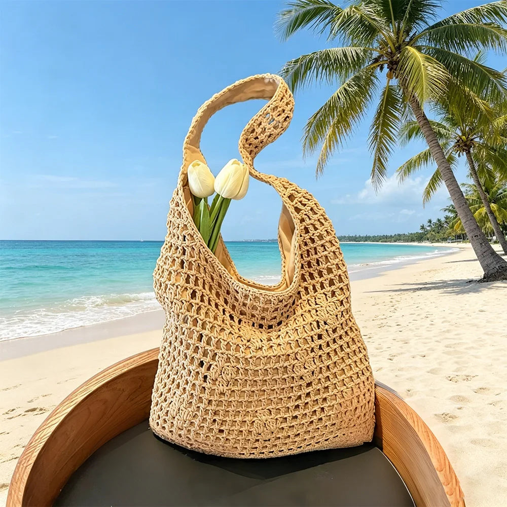 A woven crochet tote bag resting on a wooden tray on a sandy beach with palm trees and blue ocean in the background.