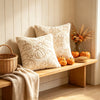 A beige plush throw pillow cover with an embossed maple leaf design rests on a wooden shelf next to decorative pumpkins and d