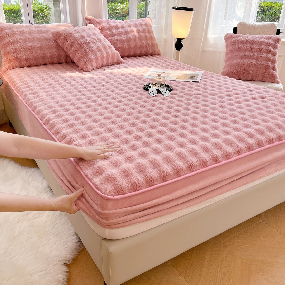 A pink luxury faux fur fitted sheet and pillowcase set displayed on a bed with white curtains.