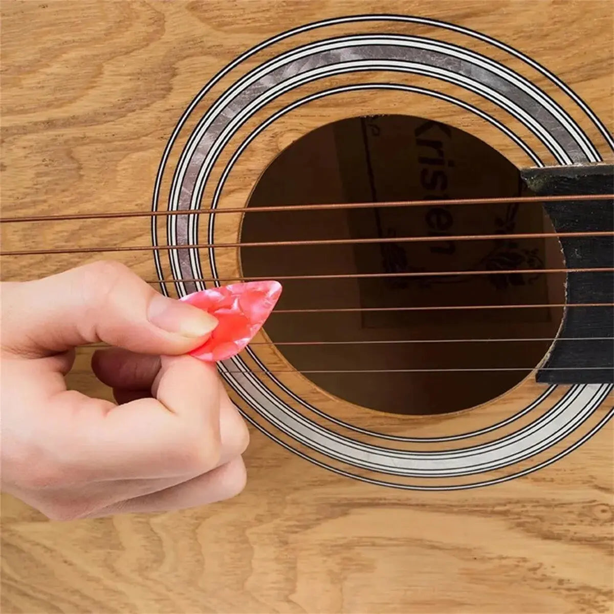 A hand is holding a pink guitar pick above an acoustic guitar fretboard.