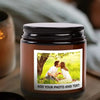 A dark brown soy candle in a glass jar with a photo and text insert, set on a wooden surface.