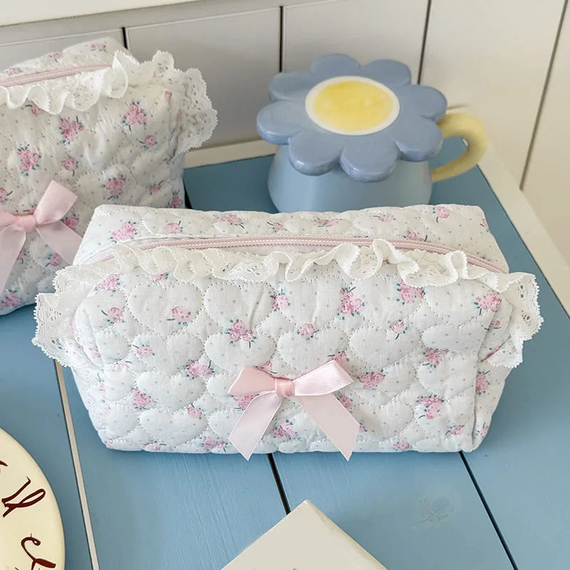 A white cosmetic pouch with a vintage rose print and lace trim is displayed next to a light blue ceramic cup.