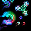 Hands manipulate various LED glow fidget spinners showing vibrant colors like blue, green, and red light.
