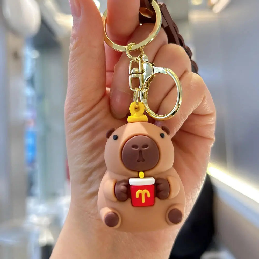 A hand holds a cute brown capybara keychain charm with a red cup attached to a gold ring.