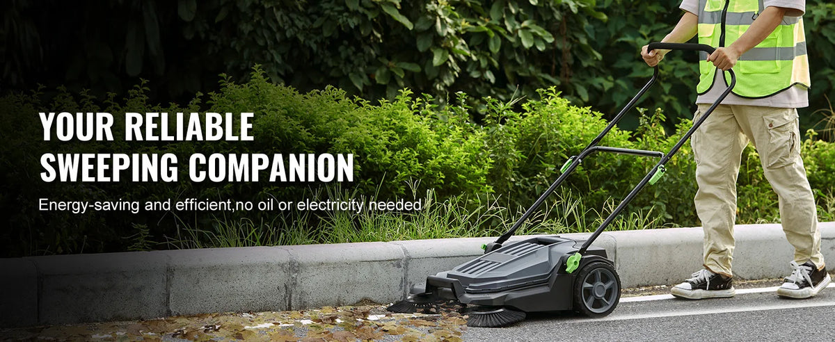 A person is operating a black walk-behind push floor sweeper outdoors on a paved surface.