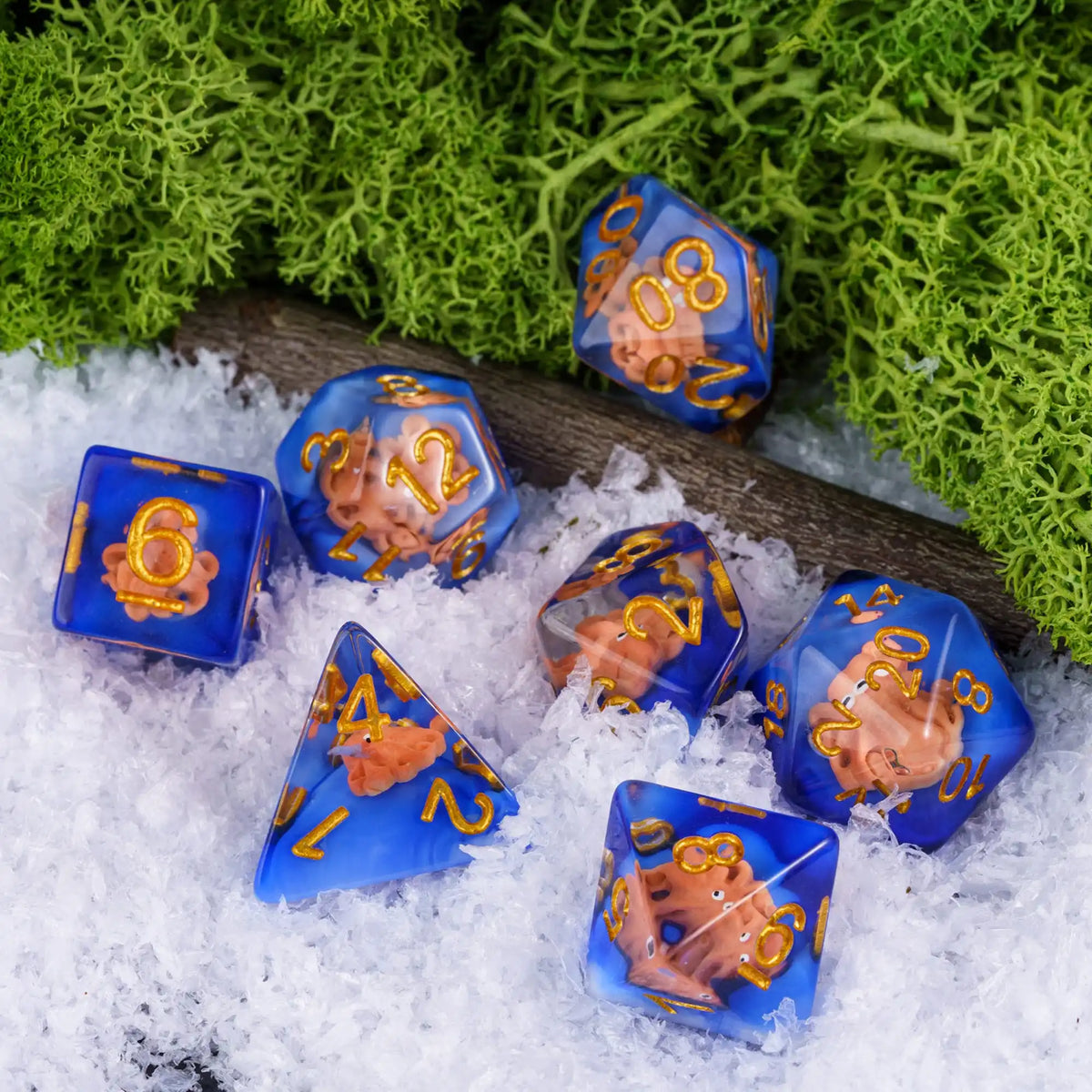 A set of adorable animal resin dice in blue and gold colors resting on white snow with green moss in the background.