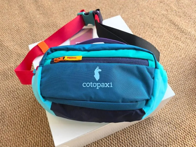 A teal waterproof nylon fanny pack with black and purple accents is shown on a white box.