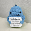 A handmade blue crochet dinosaur doll with a sad expression and a white tag that reads Small dinosaur Your future will be str