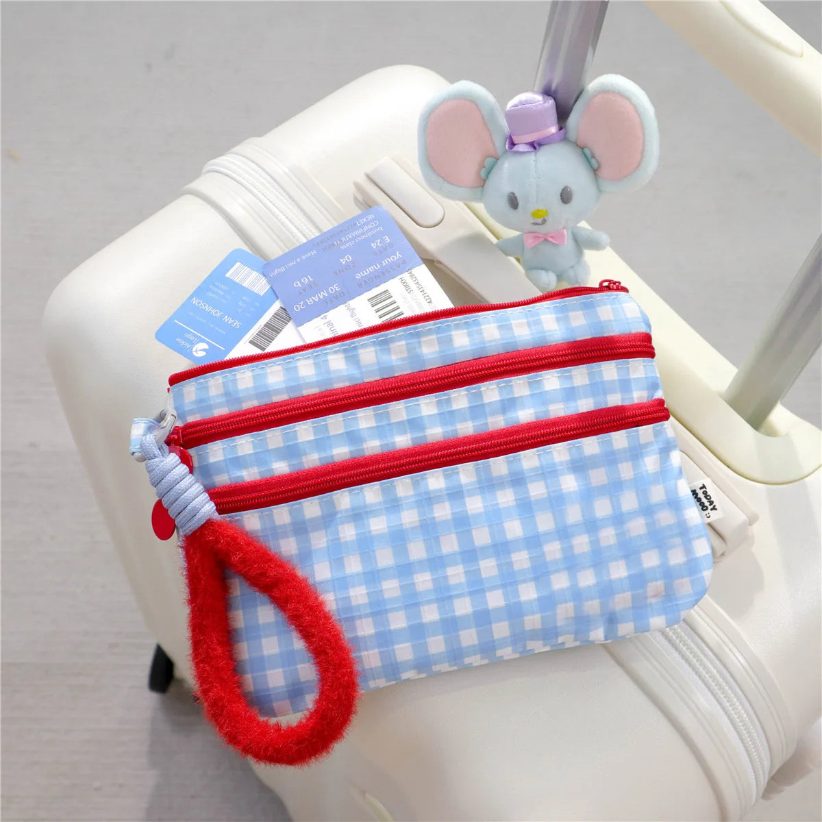A light blue plaid passport holder with a red strap and a pink elephant charm is placed on a white rolling suitcase.