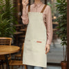 A person wearing a light pink long-sleeved shirt and a beige apron with brown straps standing indoors.