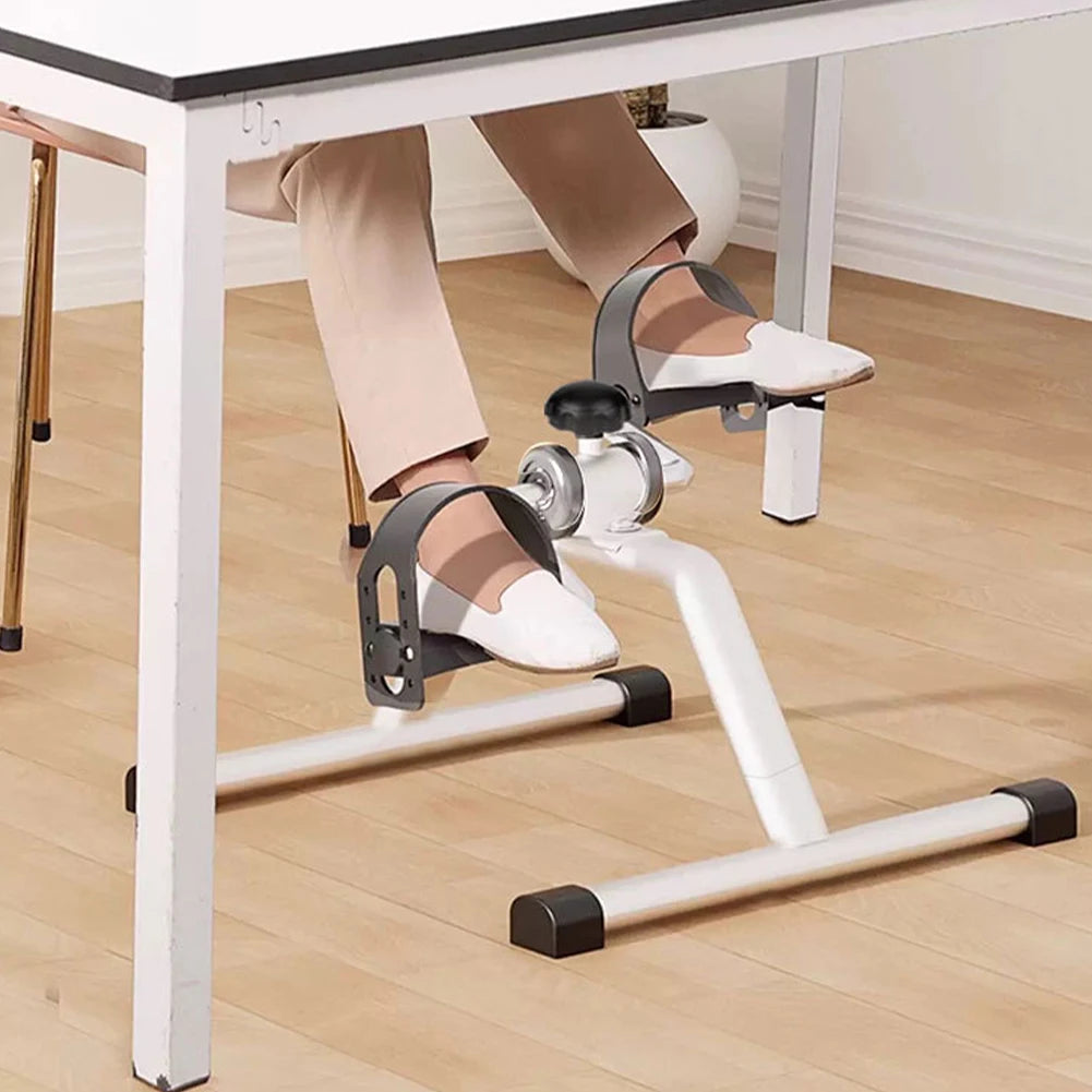 A person is using a compact under-desk pedal exerciser with adjustable resistance on a white desk.
