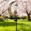 A solar moon-shaped garden light featuring an owl and a glass ball hangs from a stake in a blooming orchard.