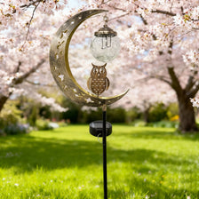 A solar moon-shaped garden light featuring an owl and a glass ball hangs from a stake in a blooming orchard.