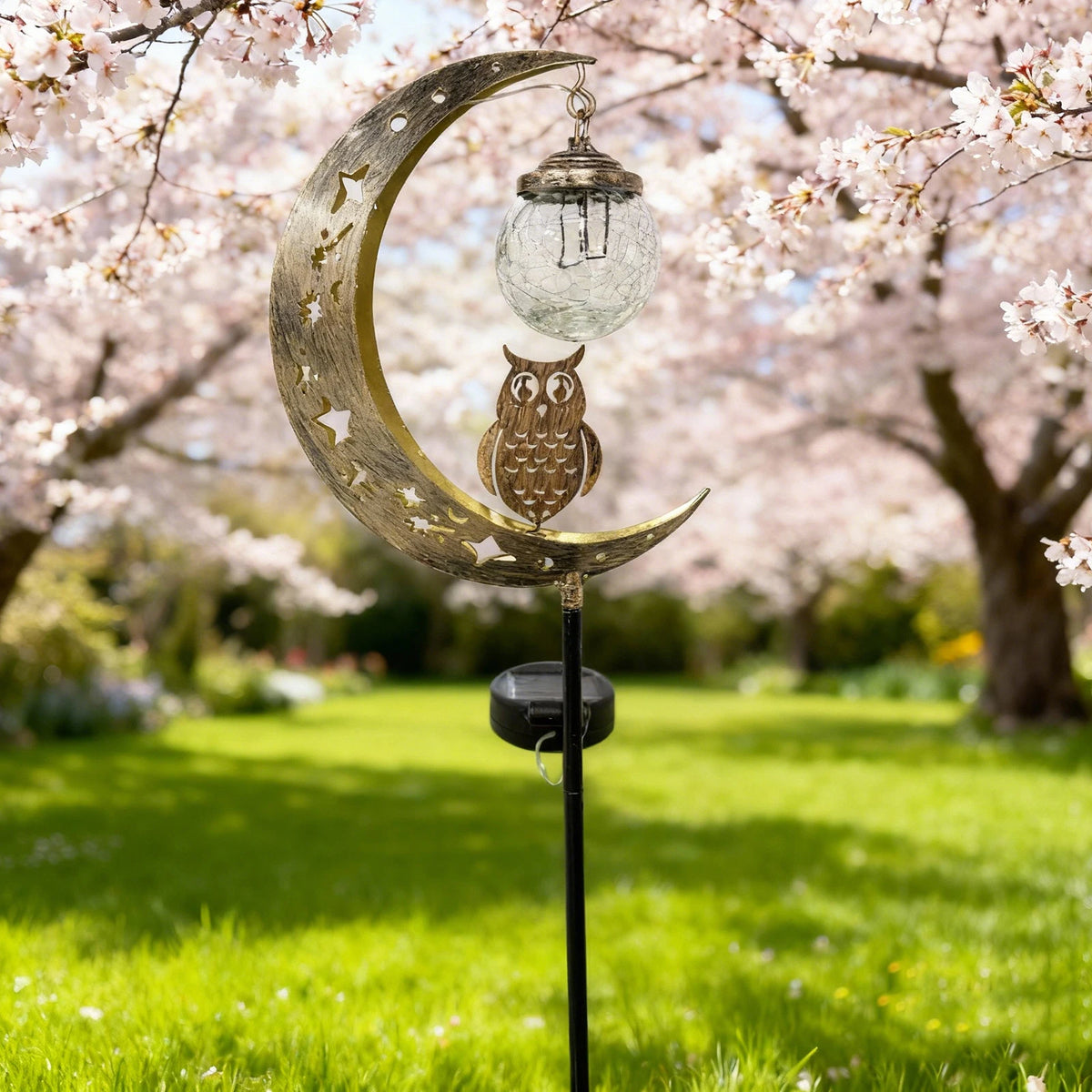 A solar moon-shaped garden light featuring an owl and a glass ball hangs from a stake in a blooming orchard.