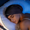 A woman wearing a black silk satin sleep bonnet, demonstrating an adjustable elastic band with a buckle.