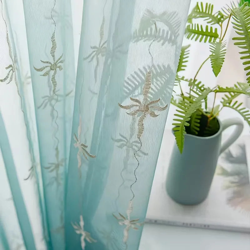Light blue sheer curtain with silver floral embroidery hanging in a window.