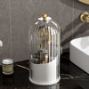 A clear glass rotating makeup brush holder with a gold lid displayed on a black marble surface.