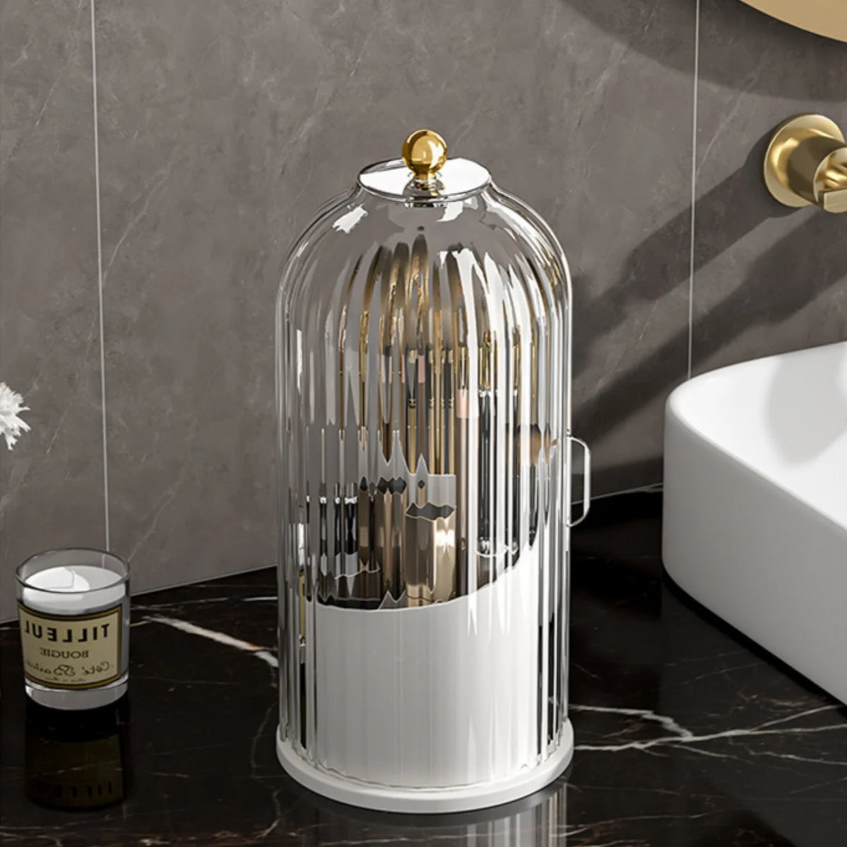 A clear glass rotating makeup brush holder with a gold lid displayed on a black marble surface.