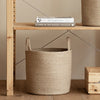 A handwoven natural fiber storage basket with handles sits on a wooden shelf next to a book.
