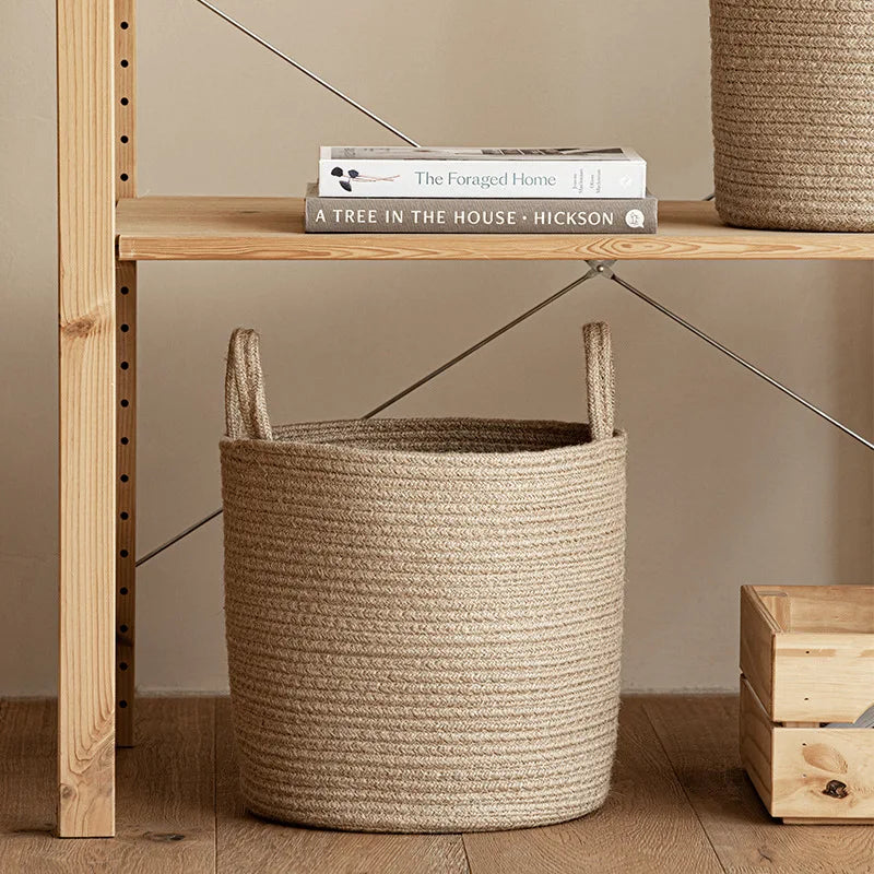A handwoven natural fiber storage basket with handles sits on a wooden shelf next to a book.