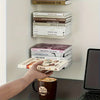 A hand holds a book near a matte black floating bookshelf set of four heavy-duty wall-mounted storage boxes.