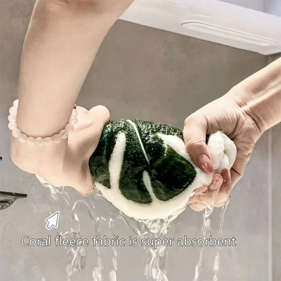 Hands are holding a green leaf-shaped coral fleece fabric under running water, demonstrating its absorbency.