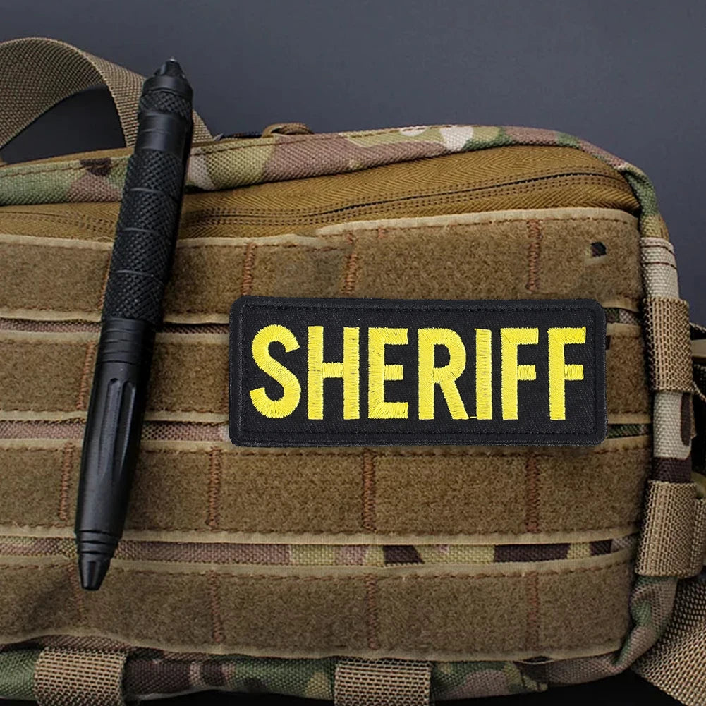 A dark green tactical backpack features a black pen and a yellow sheriff patch with hook and loop backing.