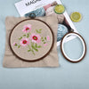 A wooden embroidery hoop with pink floral embroidery rests on a beige fabric next to yarn and citrus slices.