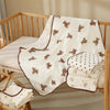 White cotton gauze baby swaddle blanket with brown bear print and decorative stitching displayed on a wooden crib.