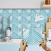 Hands holding a rectangular tile featuring a blue and white herringbone pattern against a tiled backsplash.