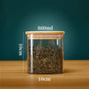 A clear glass storage canister with a bamboo lid filled with dried tea leaves on a wooden surface.