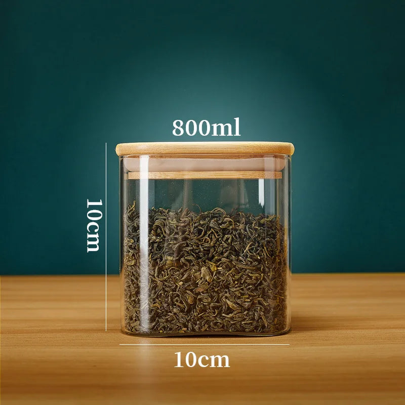 A clear glass storage canister with a bamboo lid filled with dried tea leaves on a wooden surface.