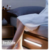 A bedside lamp illuminates a wooden nightstand next to a bed with gray pillows and light-skinned legs.