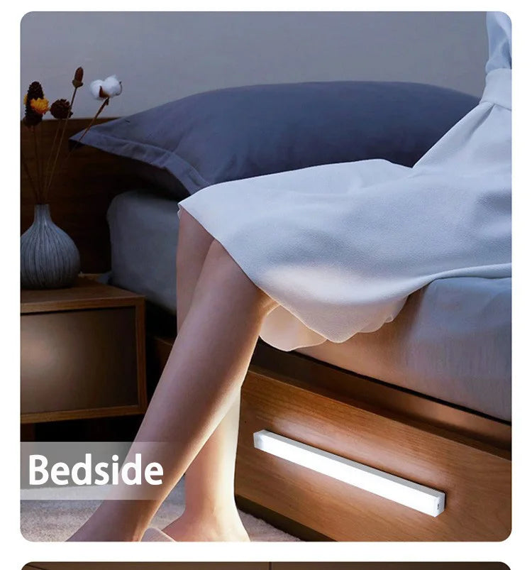 A bedside lamp illuminates a wooden nightstand next to a bed with gray pillows and light-skinned legs.