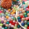 A close-up of various round imitation pearl beads in shades of pink, green, blue, and gold scattered on a white surface.