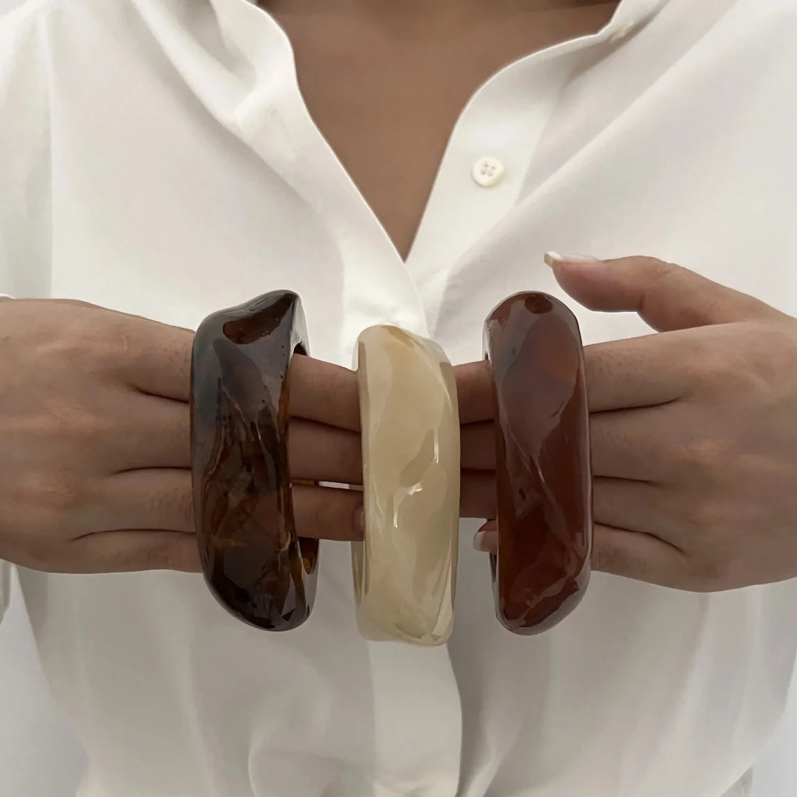 Gros plan d'une main portant trois bracelets de poignet en acrylique marron et crème sur un t-shirt blanc.