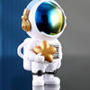 A white resin figurine of an astronaut with a reflective blue helmet and gold accents, displayed against a dark blue backgrou
