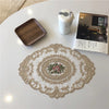 An elegant oval lace embroidered placemat with floral details rests on a white table next to a ceramic cup and a booklet.