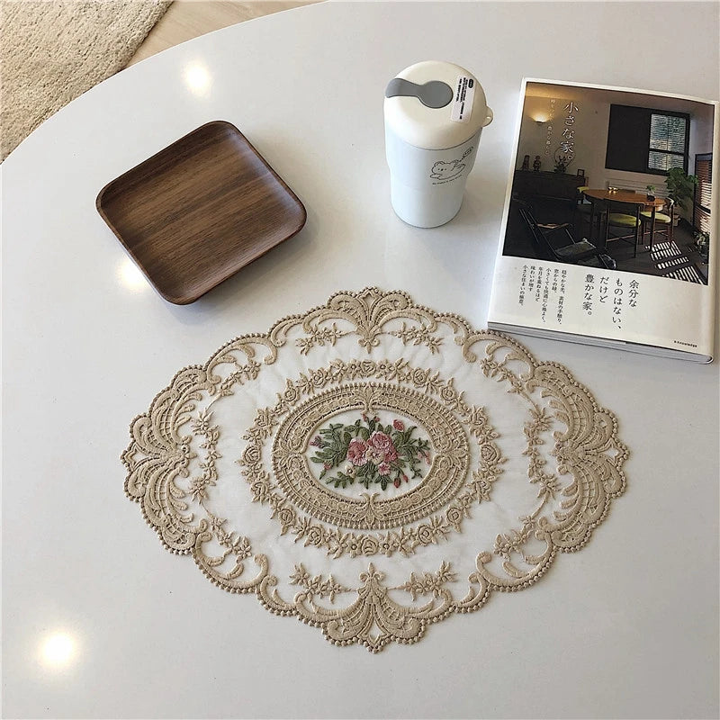 An elegant oval lace embroidered placemat with floral details rests on a white table next to a ceramic cup and a booklet.