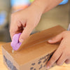 A hand is using a purple cloud-shaped retractable letter opener with a keychain loop to open a cardboard box.